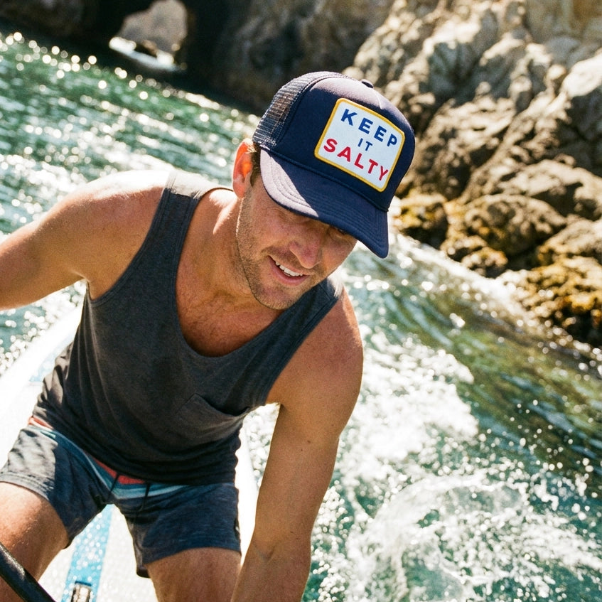 Man on a boat wearing a cap with 'Keep It Salty' text, surrounded by rocky coastline and water.