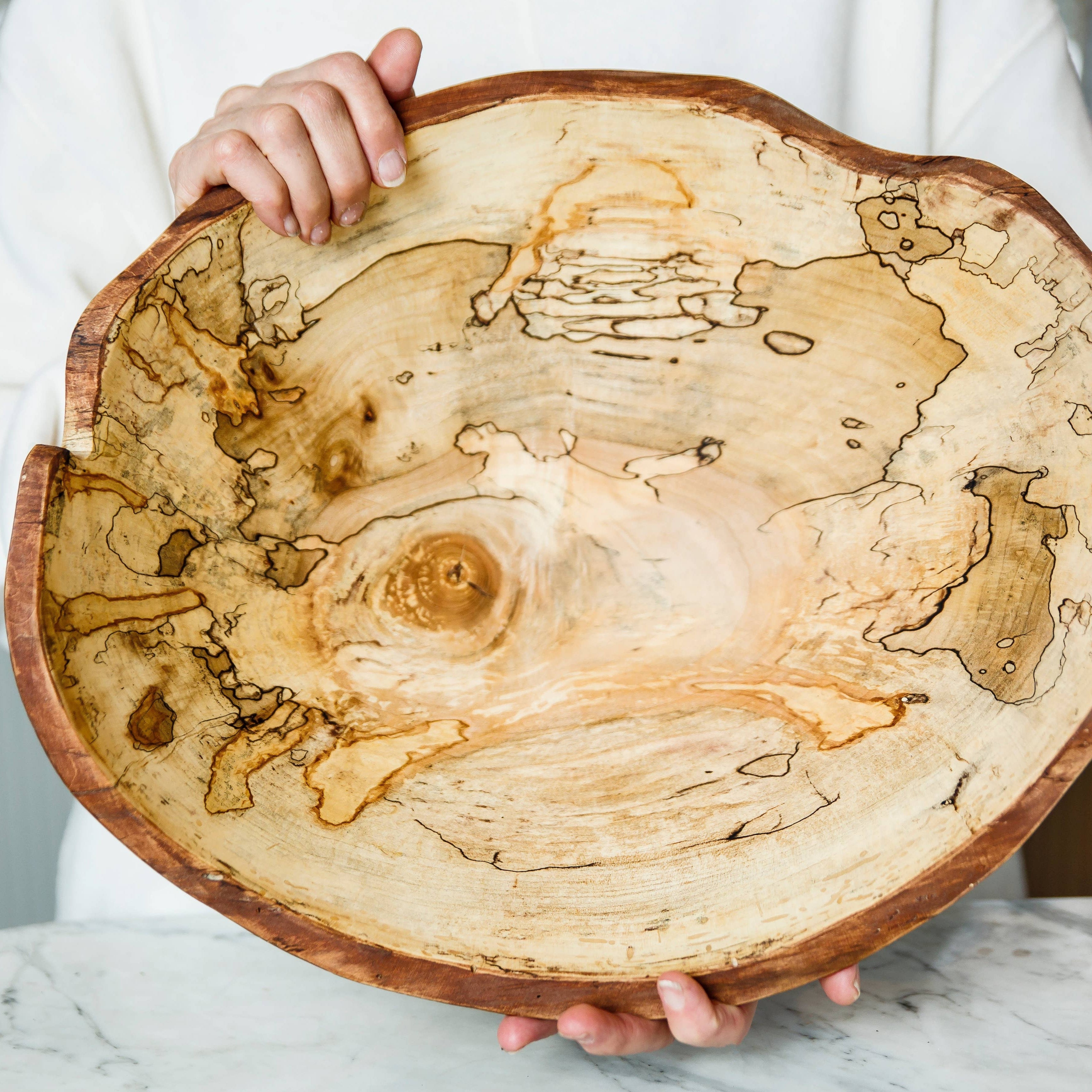 A person holding a medium-sized oval serving bowl made of spalted maple wood with a smooth finish.
