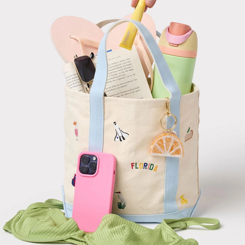 Beige tote bag with colorful handles, pink phone case, and green fabric on a light gray background