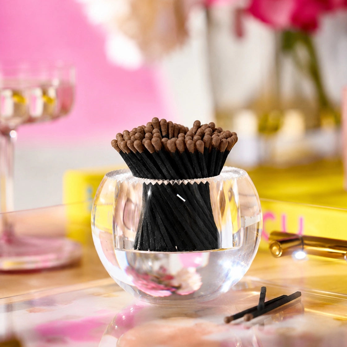 A clear glass bowl filled with black incense sticks that have brown tips, set on a reflective pink and yellow table with a blurred decorative background.