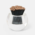A clear glass bowl-shaped holder filled with black matchsticks topped with tan wooden tips, photographed on a clean white background.