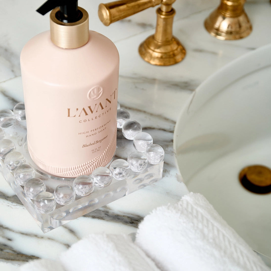 A bottle of hand wash in a pale pink bottle with a gold collar and black pump, resting on a clear glass tray on a white marble countertop next to white towels and a sink.