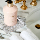 A bottle of hand wash in a pale pink bottle with a gold collar and black pump, resting on a clear glass tray on a white marble countertop next to white towels and a sink.