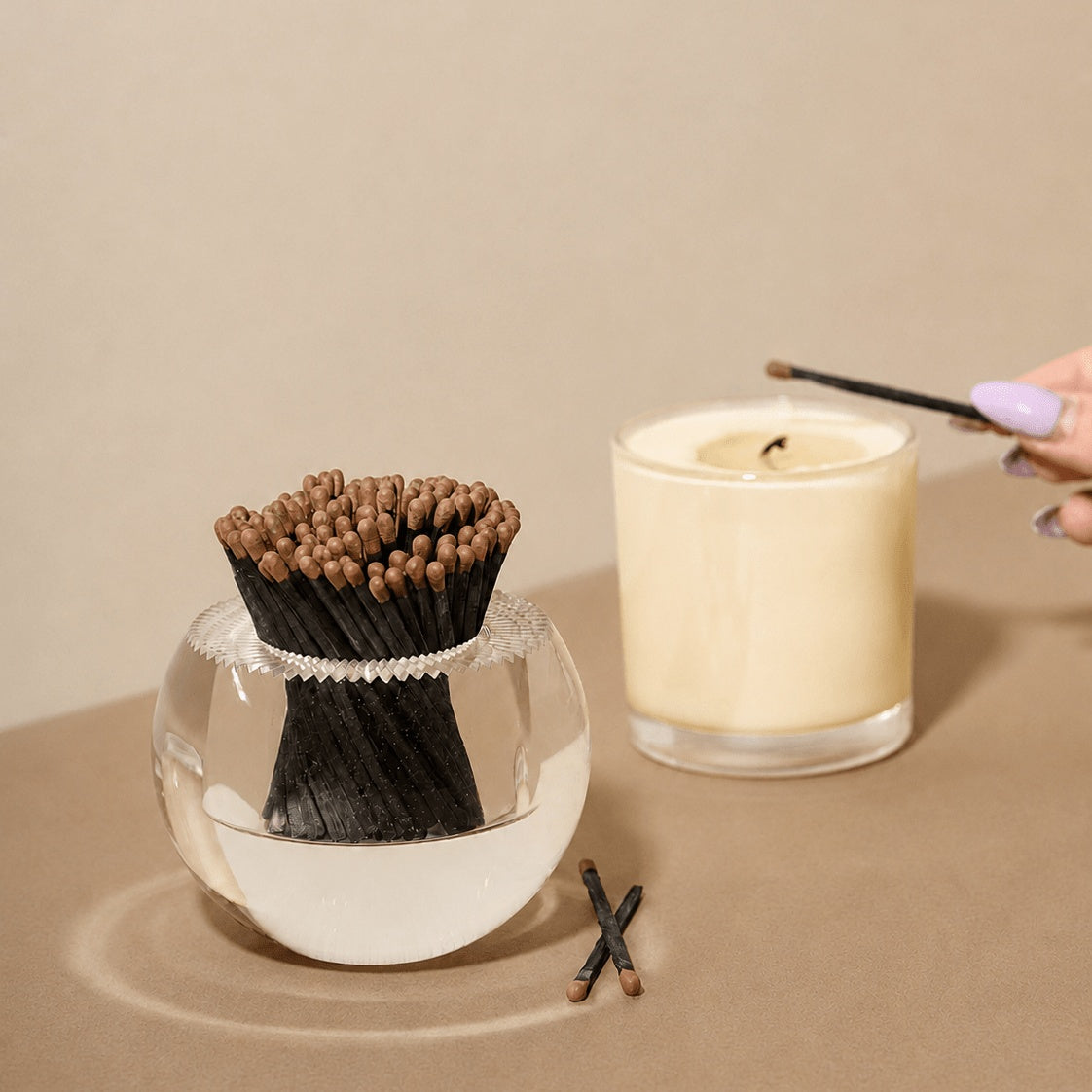 A set of black incense sticks in a clear glass bowl with a white rim, shown next to a lit cream-colored candle on a beige surface. A hand with lavender-painted nails is holding a matchstick near the candle.