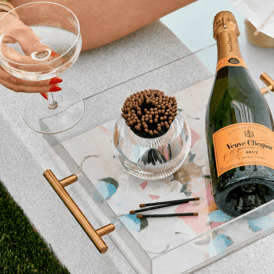 A clear acrylic cocktail tray with gold handles and a floral print, styled on a gray table outdoors. The tray holds a bottle of Veuve Clicquot Brut, a small glass bowl of brown matches, and three black cocktail sticks, with a hand holding a coupe glass visible in the upper left.