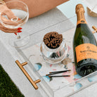 A clear acrylic cocktail tray with gold handles and a floral print, styled on a gray table outdoors. The tray holds a bottle of Veuve Clicquot Brut, a small glass bowl of brown matches, and three black cocktail sticks, with a hand holding a coupe glass visible in the upper left.
