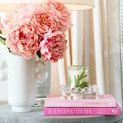 A styled interior scene with a white vase holding large pink peony flowers on a gray side table, next to a lamp and a small candle on stacked pink books.